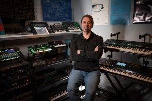 A white man in a sweater, jeans, and knit hat sits with his arms crossed among keyboards and music recording equipment.